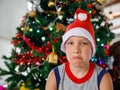 Portrait of upset and sad boy in red Santa hat sits in front of Christmas tree and angrily looks at camera Royalty Free Stock Photo