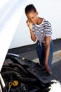 Upset young african woman standing by broken down car parked on the side of a road and calling for assistance Royalty Free Stock Photo