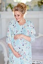 The portrait is very beautiful, mile, feminine and tender pregnant girl blonde in blue dress with floral print against a light in Royalty Free Stock Photo