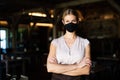 Waitress with face mask standing indoors on terrace restaurant, arms crossed.