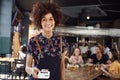 Portrait Of Waitress Holding Credit Card Payment Terminal In Busy Bar Restaurant