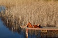 Portrait of a woman in autumn cozy orange clothes with backpack sitting on a wooden bridge near the lake with blue water Royalty Free Stock Photo