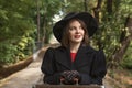 Portrait woman in black hat with brims, classic jacket and gloves rests hands on old suitcase. Forest background