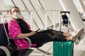 Portrait of woman black trousers, pink cardigan, white disposable mask, sitting, stretching legs, holding smart phone. Royalty Free Stock Photo