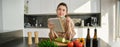 Portrait of woman checking recipe notes in notebook, standing in kitchen with vegetables, cooking food, preparing Royalty Free Stock Photo