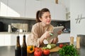 Portrait of woman checking recipe notes in notebook, standing in kitchen with vegetables, cooking food, preparing Royalty Free Stock Photo