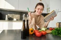 Portrait of woman checking recipe notes in notebook, standing in kitchen with vegetables, cooking food, preparing Royalty Free Stock Photo