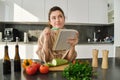 Portrait of woman checking recipe notes in notebook, standing in kitchen with vegetables, cooking food, preparing Royalty Free Stock Photo