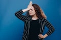 Portrait of woman with curly dark hair forgetting something, slapping forehead with palm and closing eyes over blue