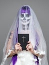 Portrait of woman looks at the camera with terrifying halloween skeleton makeup and purple wig bridal veil, wedding dress, holds t