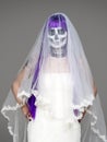 Portrait of woman looks at the camera with terrifying halloween skeleton makeup and purple wig bridal veil, wedding dress over gra