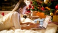 Portrait of young woman lying under Christmas tree and doing online shopping on laptop Royalty Free Stock Photo