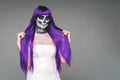 Portrait of woman with terrifying halloween skeleton makeup and purple wig hair and wedding dress over gray background looks at