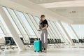 Portrait of woman wearing black T-shirt, grey trousers, white disposable mask, standing, holding turquoise suitcase. Royalty Free Stock Photo