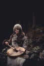 Portrait of a woman wearing fur hat and leather jacket.Playing shamanic drum in the forest.