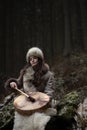 Portrait of a woman wearing fur hat and leather jacket.Playing shamanic drum in the forest.