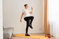 Portrait of woman wearing white t shirt and black leggins doing sport exercises at home, standing on mat and matching, looking Royalty Free Stock Photo