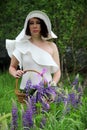 Portrait of a woman in a white hat with a basket of lilac lupines, vertical photo. A brunette in a white dress and a