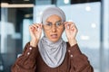 Portrait of worried young muslim woman in hijab sitting in office and looking surprised at camera holding glasses with Royalty Free Stock Photo