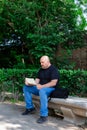 Portrait of a 45-50 year old bald man sitting alone on an outdoor park bench on a sunny afternoon reading a book.