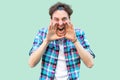 Portrait of yelling young man in casual blue checkered shirt and headband standing with hands on his face, shouting or screaming Royalty Free Stock Photo