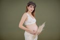 Portrait of young adorable pregnant woman with long dark hair wearing brown hat holding belly and dried pampas grass.