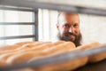 Portrait of young adult baker with long beard in white uniform standing in his workplace and looking on fresh loaf in the tray Royalty Free Stock Photo