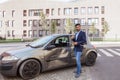 Portrait of young adult bearded insurance agent standing with positive facial expression near wrecked car and showing thumb up,