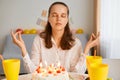 Portrait of young adult Caucasian dark haired female in white shirt sitting at table with birthday cake, celebrating party alone,
