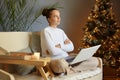Portrait of young adult caucasian girl sitting on sofa with crossed legs working using laptop by christmas tree, thinking about Royalty Free Stock Photo