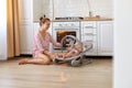 Portrait of young adult Caucasian mother wearing casual striped shirt and short sits on floor with toddler daughter in rocking Royalty Free Stock Photo