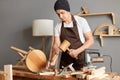 Portrait of young adult man carpenter wearing white t-shirt, black cap and brown apron, craftsman carve with a gouge in the hands Royalty Free Stock Photo