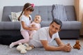 Portrait of young adult man lying on floor near sofa and playing with his little kids, smiling kids sitting on dad`s back, Royalty Free Stock Photo