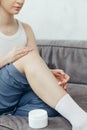 Portrait of young adult woman applying body lotion for skin on legs while sitting on couch at home Royalty Free Stock Photo