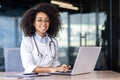 Portrait of a young African American female doctor in a white coat sitting at a table and working at a laptop, looking Royalty Free Stock Photo