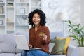 Portrait of a young African American woman sitting on the couch at home, holding a laptop on her lap and a credit card Royalty Free Stock Photo