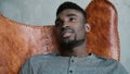 Portrait of young African male sitting in leather chair, thinking about something and smiling. Man looks thoughtfully. Royalty Free Stock Photo