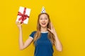 Portrait of young amazed woman holding in hand gift having fun, standing in a birthday cone and blue casual t shirt over yellow