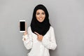 Portrait of young arab woman 20s in islamic headscarf with oriental makeup pointing finger on black screen of cell phone, isolate Royalty Free Stock Photo