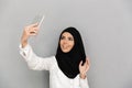 Portrait of young arab woman 20s in islamic headscarf with oriental makeup taking selfie on silver smartphone, isolated over gray Royalty Free Stock Photo