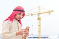 Portrait of young arabian holding the smartphone and looking to mobile screen at construction site with crane background