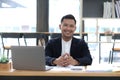 Portrait of a young Asian businessman smiling while using a laptop and writing down notes while sitting at his desk in a Royalty Free Stock Photo