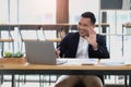 Portrait of a young Asian businessman smiling while using a laptop and writing down notes while sitting at his desk in a Royalty Free Stock Photo