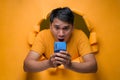 Portrait Young Asian man through torn yellow paper hole, astonished looking at smartphone screen in shock Royalty Free Stock Photo