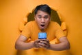 Portrait Young Asian man through torn yellow paper hole, stonished looking at smartphone screen in shock Royalty Free Stock Photo