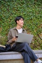 A young Asian man remote working in the city garden park, using laptop, sitting on a bench Royalty Free Stock Photo