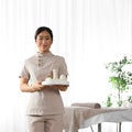 Portrait of young Asian masseuse holding tray with spa stuff in salon