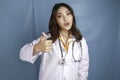 Portrait of a young Asian woman doctor, a medical professional is smiling and showing thumbs up or OK sign isolated over blue Royalty Free Stock Photo