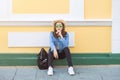 Portrait young Asian woman wearing sunglass in jean jacket and straw hat sitting on floor touch her chin nextt yellow wall