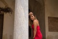 Portrait of young, attractive, blonde woman in an elegant red party dress leaning on a marble column smiling happily. Concept Royalty Free Stock Photo